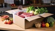 © Iona - A box containing fresh meat and various vegetables is open on a wooden kitchen countertop, ready for meal preparation.