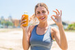 © luismolinero - Young pretty sport woman holding an orange juice at outdoors showing ok sign with fingers