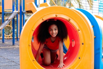 Young Afro hair elementary schoolgirl crawling through colorful tunnel on a playground, weekend carefree holiday in park. bright playground equipment and focus expression capture childhood exploration