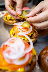 Wall Mural - A close-up of hands preparing a gourmet burger, adding pickles on top of fresh ingredients like lettuce, tomato, and cheese. Perfect for culinary themes, food blogs, or restaurant promotions.