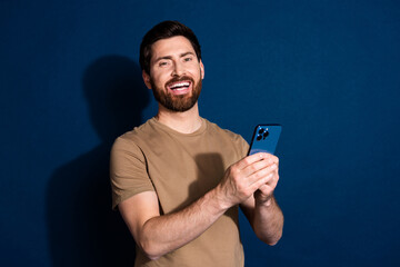 Wall Mural - Photo portrait of young laughing guy brunet hair model in beige t shirt using smartphone read memes isolated on dark blue color background