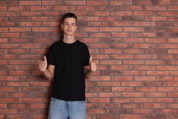 Canvas Print - Teenage boy wearing black t-shirt and showing thumbs up near brick wall