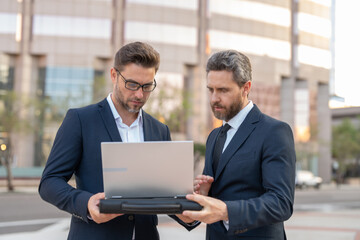 Two businessman on the street, deeply engrossed, eyes fixed on their laptop screen. Two handsome young businessmen in classic suits using laptop. Business men team using laptop outdoor. Business man