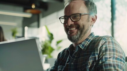 Sticker - A smiling man in glasses and a checkered shirt, working on his laptop in a bright, modern office with greenery in the background.