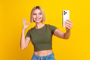 Wall Mural - Photo of positive girl with bob hairdo dressed khaki crop top hold smartphone show okey make selfie isolated on yellow color background