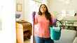 © AntonioDiaz - Plus size Hispanic woman stands in a home living room, holding a mop and a bucket of cleaning supplies