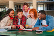 © Jose Calsina - Diverse group of college teenage students studying together, looking at notes and using a cellphone while collaborating at university campus library in a cheerful, casual setting. Five people teamwork
