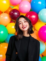 Sticker - A joyful Asian woman with short black hair smiles broadly while wearing a stylish black blazer. The background is a vibrant and festive wall of colorful balloons, creating a celebratory atmosphere.