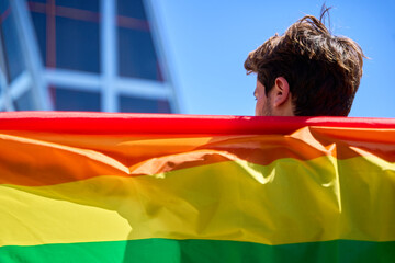 A man holding a rainbow flag in front of a building. The flag is large and colorful.