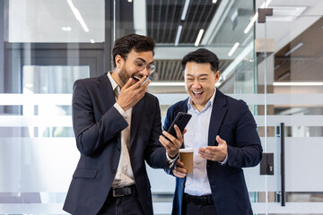 Two colleagues sharing a light moment in office while watching a funny video on smartphone. Both men dressed in business suits, one holding a coffee cup, expressing laughter and surprise.