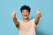 © SHOTPRIME STUDIO - Happy young man with curly hair giving two thumbs up, smiling joyfully against a light blue background, embodying positivity and enthusiasm in a casual setting