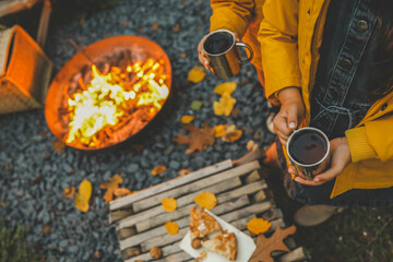 Children in warm clothing holding cups of hot drink near a bonfire in the evening. Cozy autumn outdoor scene with a fire pit, fallen leaves, and rustic snacks, capturing the essence of fall warmth