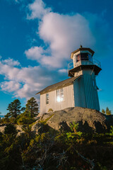 Close-up view of a historic wooden lighthouse standing on rugged rocky terrain, illuminated by sunlight and set against a vibrant blue sky with clouds. Ideal for maritime, heritage, and nature themes.