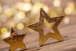 © drubig-photo - Two golden star-shaped ornaments with a rustic texture on a wooden surface, with a blurred background featuring warm bokeh lights, creating a festive and cozy atmosphere.
