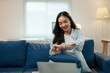 © amnaj - Young woman is sitting on a blue sofa and checking the time on her smartwatch while working remotely on her laptop