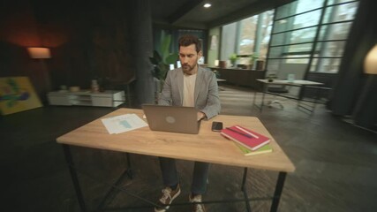 Footage of programmer working on laptop in modern cozy office. Young Caucasian man sitting at working place. Camera moving around. 360 degree tracking shot. Parallax effect. Speed