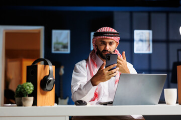 A male entrepreneur, dressed in contemporary clothing, using a digital laptop and mobile device, confidently speaking on phone call in a modern office, symbolizing success in the digital age.