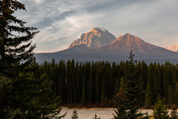 view of a valley in the mountains with a coniferous forest under the first rays of sunshine and orange light on the mountain peak and light clouds in the sky