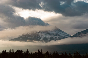 A breathtaking view of majestic snow-covered mountains partially veiled by clouds and fog, creating a serene and mystical atmosphere over the wooded landscape.