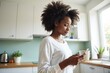 © Derek Brumby - A woman stands in her modern kitchen, holding a cup of coffee in one hand and checking her smartphone with the other, representing a blend of relaxation and connectivity.