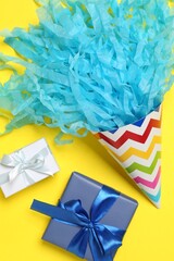 Wall Mural - Party hat with beautiful tissue paper and gift boxes on yellow background, flat lay