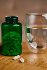 Close-up of two white pills near green bottle with glass of water on wooden table. Concept of vitamins and supplements for health care