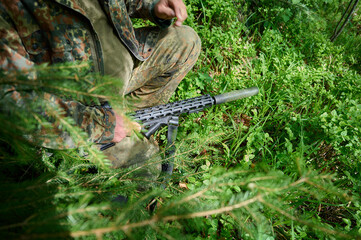 Hunter with rifle wearing camouflage suit sitting in ambush. Camouflaged sniper in the forest in ambush. Military man hide in the woods with rifle.