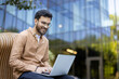 © Liubomir - Young man smiling while using laptop outdoors in modern office environment demonstrating work flexibility and remote productivity. Business attire suggests professionalism and confidence