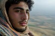 © Outkast - Close-up Portrait of a Young Man with Curly Hair and a Goatee, Wearing a Patterned Scarf, Looking Away, Against a Blurry Landscape Background