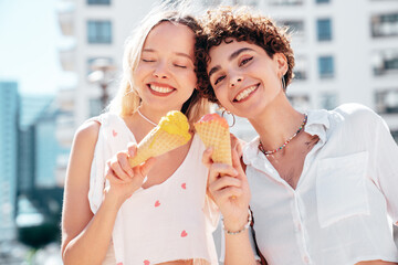 Two young beautiful smiling hipster female in trendy summer clothes. Carefree women posing on street background. Positive models eating tasty ice cream in waffles cone in sunny day, cheerful and happy