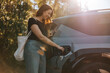 © Maskot - Smiling mature woman charging car while standing at station