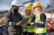 © TATIANA Z - Construction workers wearing safety helmets and vests, discussing project details using a tablet at an outdoor industrial site