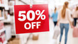 © Anna - 50% off sale sign hanging in busy retail store with shoppers in background.
