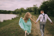 © Vasil - A couple is running together in a grassy field near a lake. The woman is wearing a green dress and the man is wearing a white shirt and black suspenders. Scene is joyful and romantic