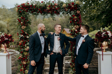 Wall Mural - Three men in suits stand under a red archway, posing for a photo. The archway is decorated with flowers, and there are vases on the ground. Scene is celebratory and festive