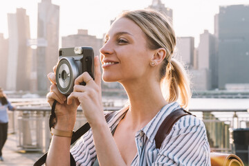 Wall Mural - Carefree female tourist with stylish rucksack standing at urban setting with Manhattan district on background and focusing lens of instant camera for clicking pictures, concept of recreating