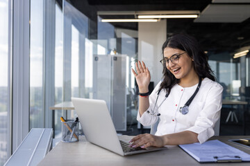 Wall Mural - Female doctor engaging in virtual consultation, smiling waving at laptop. Wears stethoscope, suggesting medical context. Office space creates professional atmosphere. Concept of remote healthcare