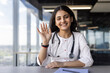 © Liubomir - Young female doctor smiling at camera, waving hand while sitting at desk. Stethoscope around neck indicates professionalism. Bright window office setting suggests modern healthcare environment.