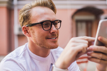 Wall Mural - Happy man in optical eyewear looking at smartphone screen and reading funny information on public website for millennials, positive male blogger messaging with virtual friends using 4g wireless