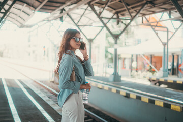 A stylish woman in sunglasses is on a phone call at a busy, vibrant train station