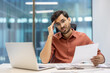 © Liubomir - Stressed man in office holding document showing confusion and worry, sitting at desk with open laptop. Business professional experiencing stress while dealing with paperwork in modern workplace.