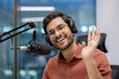 © Liubomir - Smiling man in studio speaks into microphone wearing headphones, creating podcast. Friendly wave towards camera suggests engaging interaction.