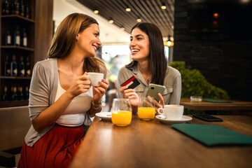 two girl best friends met at the cafe to shop online together. they are using mobile phone and credi