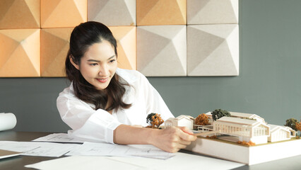 Portrait of young beautiful architect engineer measuring house model by using pencil while taking a note with architectural model, laptop and blueprint. Creative business design concept. Manipulator.