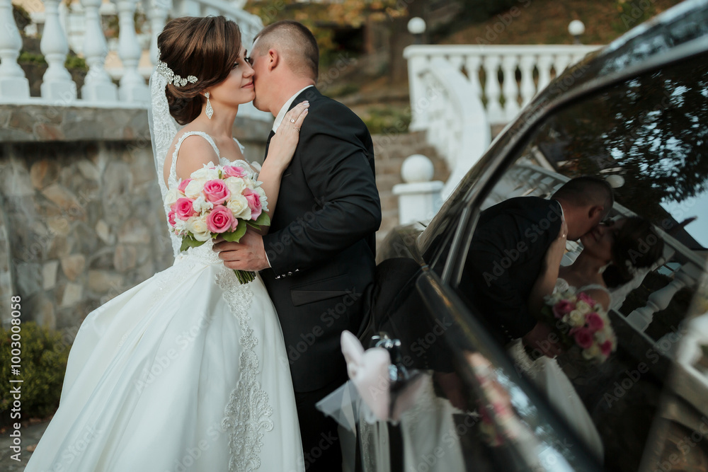 A bride and groom kiss in front of a car. The bride is wearing a white ...