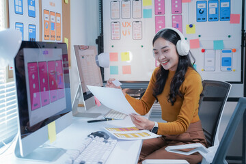 Young asian woman is working as a user experience designer in her office, designing mobile apps. She is wearing headphones and comparing printed designs