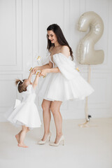 Hispanic woman and young girl in white dresses celebrating second birthday. Joyful interaction between mother and daughter with large number two balloon.