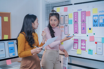 Two web designers are discussing user interface design for a mobile application, standing in their office in front of a whiteboard covered with sketches