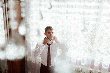 Wall Mural - A man is getting ready for a wedding, putting on a tie and adjusting his collar. The scene is set in a room with a window, and the man is looking out of it. Scene is one of preparation