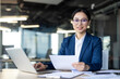 © Liubomir - Confident Asian businesswoman in formal attire at office desk holding documents with laptop. Professional setting reflects focus, success, and determination in business environment.